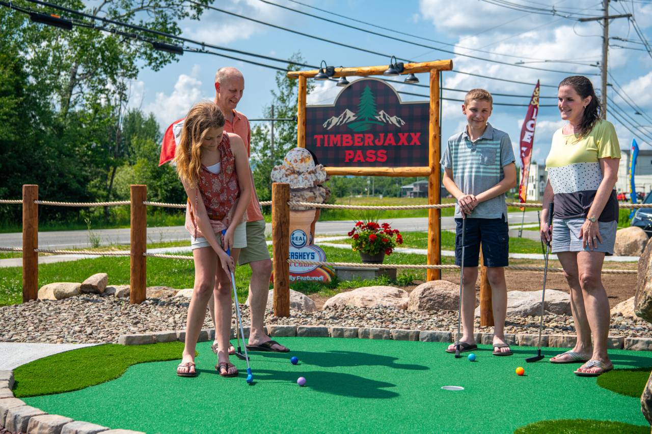 A family plays mini golf in front of a sign that reads "Timberjaxx Pass" 
